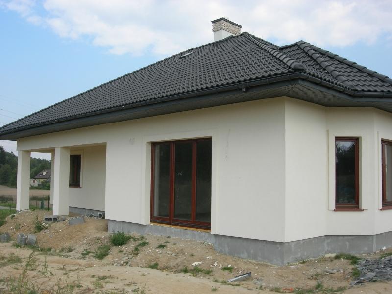 Wykończony dom jednorodzinny z ciemną dachówką i jasną elewacją. Widoczne okna z brązowymi ramami. Teren wokół domu częściowo uporządkowany.