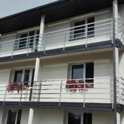 Dwupiętrowy budynek z balkonami zabezpieczonymi metalowymi barierkami z poziomych prętów, ozdobiony czerwonymi kwiatami w donicach okiennych.