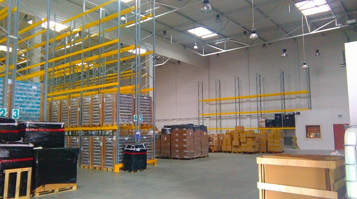 Wnętrze hali magazynowej z wysokimi regałami paletowymi, częściowo załadowanymi towarem, widoczne numery regałów i oświetlenie sufitowe.