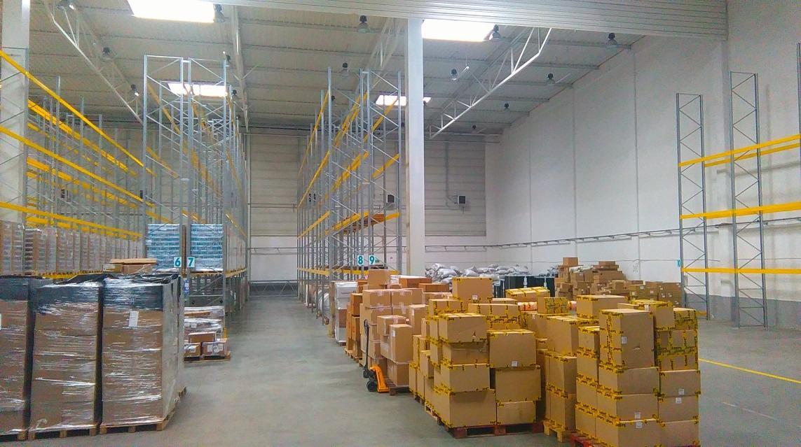 Wnętrze hali magazynowej z wysokimi regałami, paletami z kartonami i folią stretch, oświetlone światłem dziennym z okien dachowych.