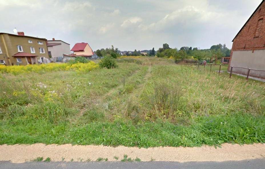 Niezagospodarowana działka budowlana w Imielinie z widocznymi śladami przejazdu pojazdów przez porośniętą trawą i chwastami powierzchnię, otoczona zabudową jednorodzinną i ogrodzeniem z siatki.