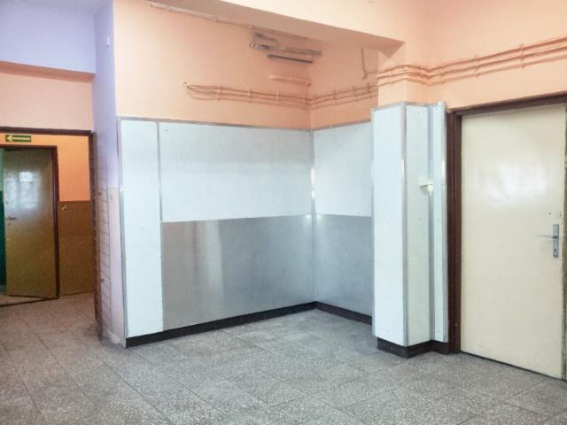 Wnętrze lokalu użytkowego z szarą podłogą z płytek, biało-szarymi ścianami i dwojgiem drzwi, z których jedne są otwarte i widać zielony znak ewakuacyjny.