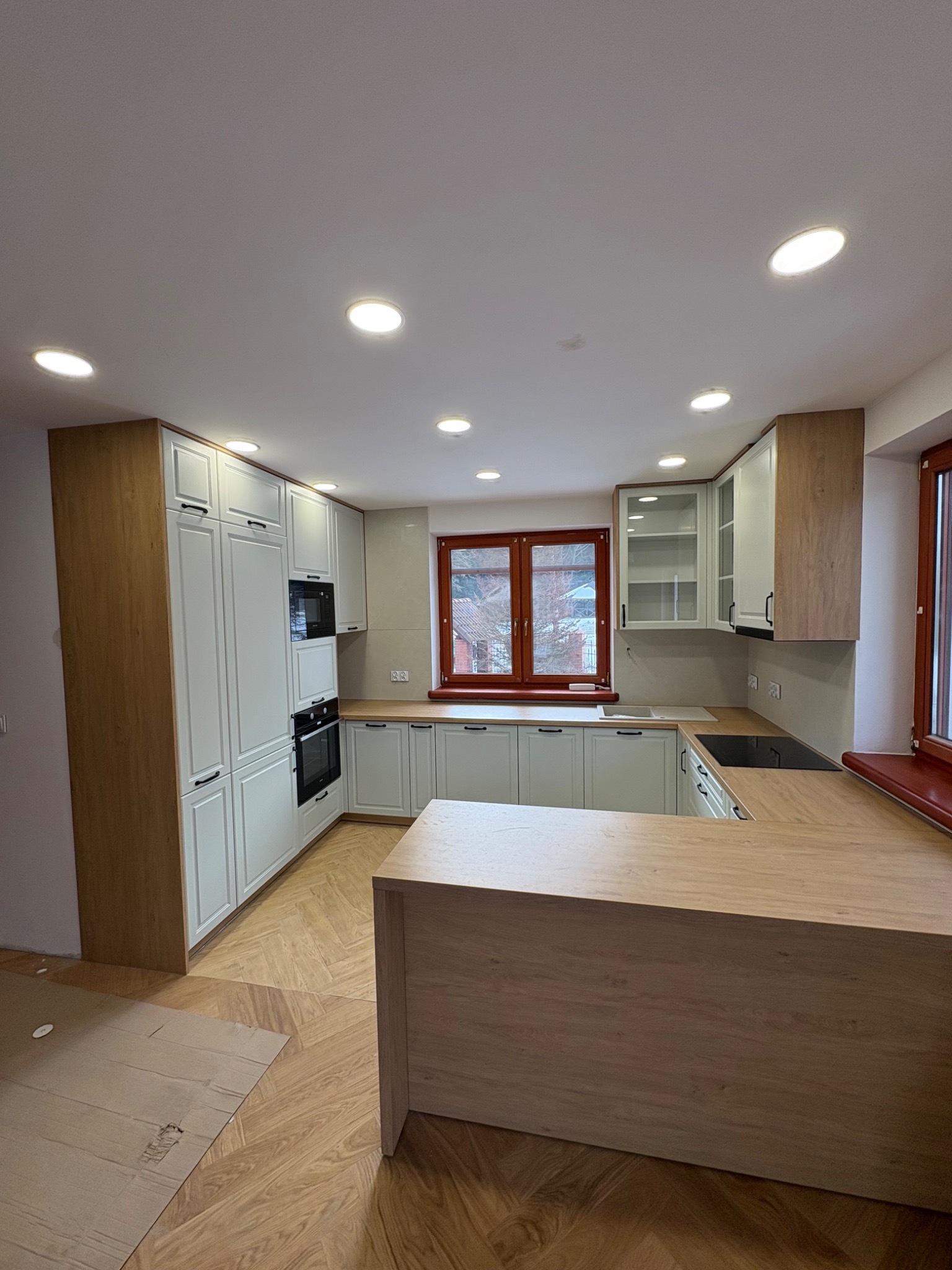 Jasna kuchnia z drewnianymi akcentami i wyspą w Zagórzu. Fronty szafek frezowane, okna z drewnianą stolarką. Parkiet ułożony w jodełkę. Nowoczesne oświetlenie.