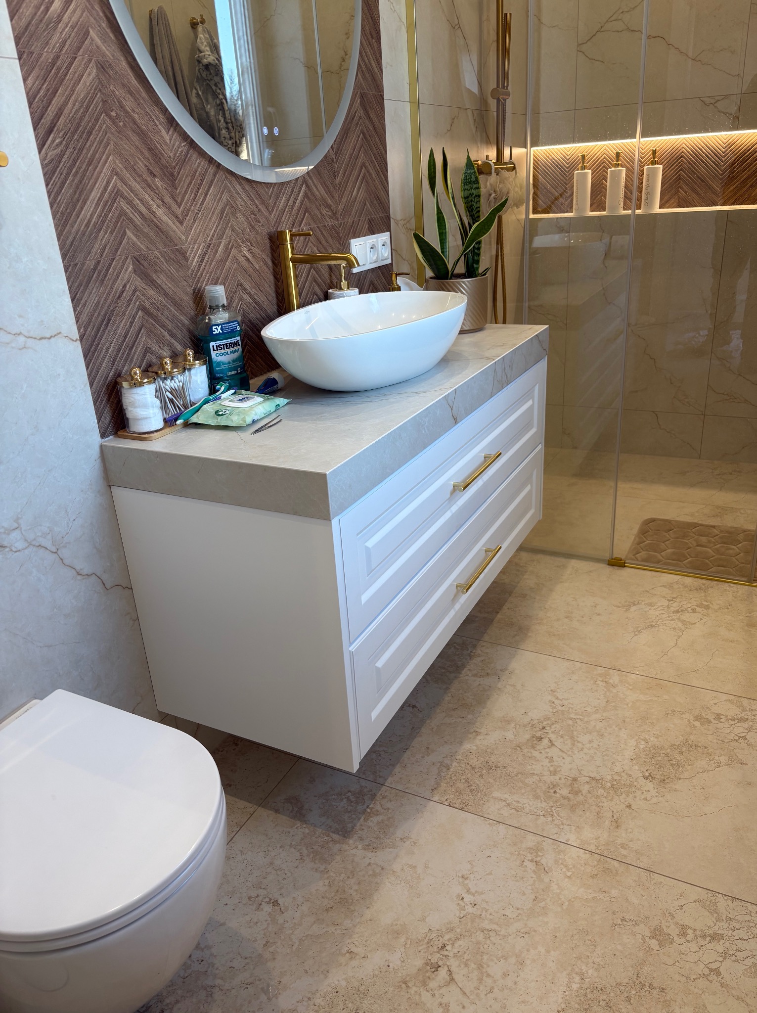Nowoczesna łazienka z białą szafką pod umywalkę z szufladami i złotymi uchwytami, owalnym lustrem i dekoracyjną ścianą w brązowe jodełki.