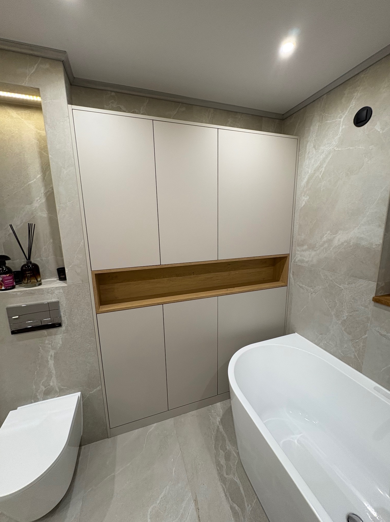 Biała szafa wnękowa z drewnianą półką w nowoczesnej łazience z wanną i toaletą. Marmurowe płytki na ścianach i podłodze. Minimalistyczny design.