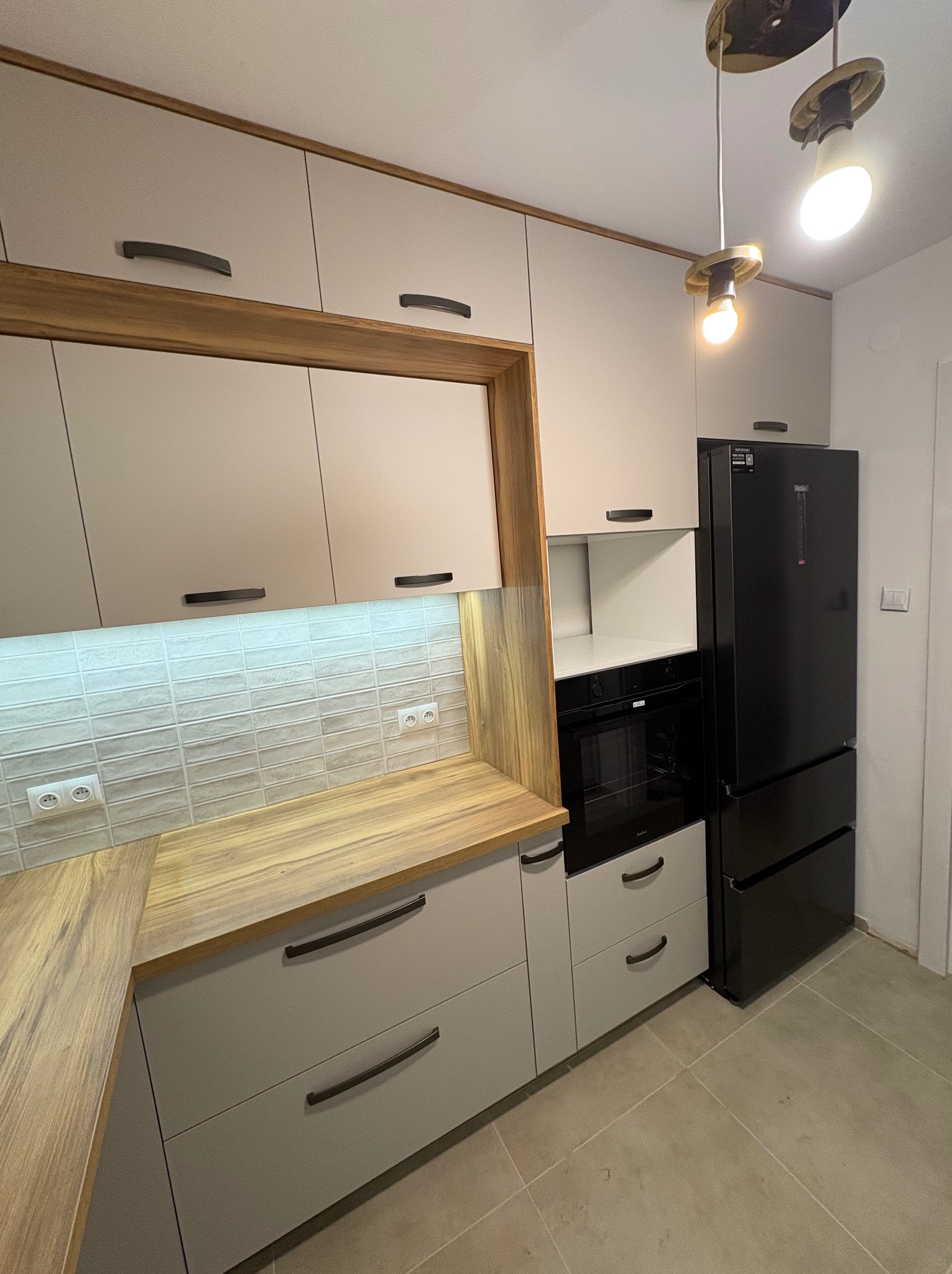 Jasna kuchnia z drewnianym blatem i czarną lodówką. Górne szafki z oświetleniem LED. Minimalistyczne uchwyty. Nowoczesne lampy sufitowe.