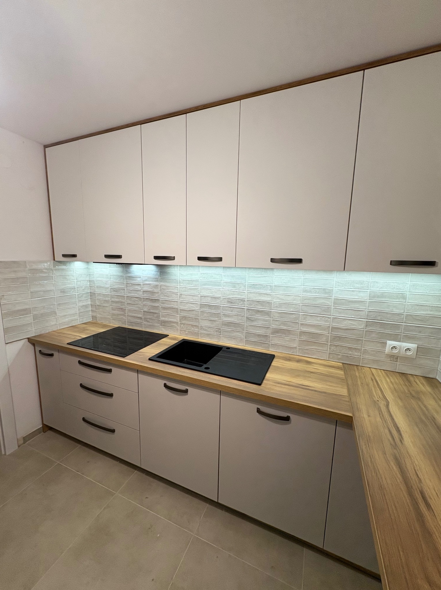 Jasna kuchnia z drewnianym blatem, czarnym zlewem i płytą. Górne szafki z oświetleniem LED. Minimalistyczny design z podłużnymi uchwytami. Płytki imitujące cegłę.