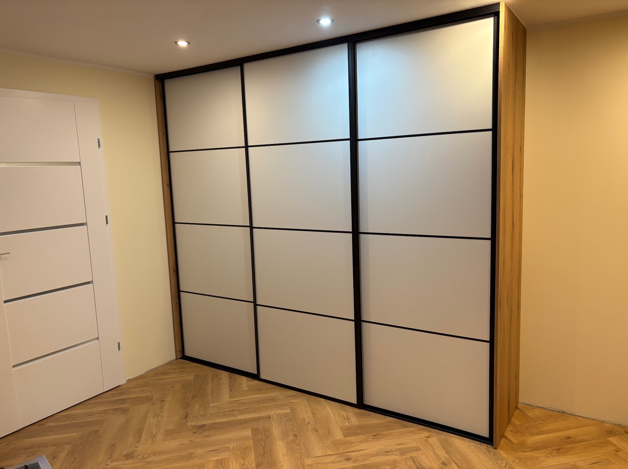 Nowoczesna szafa wnękowa z przesuwnymi, matowymi drzwiami i czarnymi ramami, wkomponowana w jasne wnętrze z drewnianą podłogą.