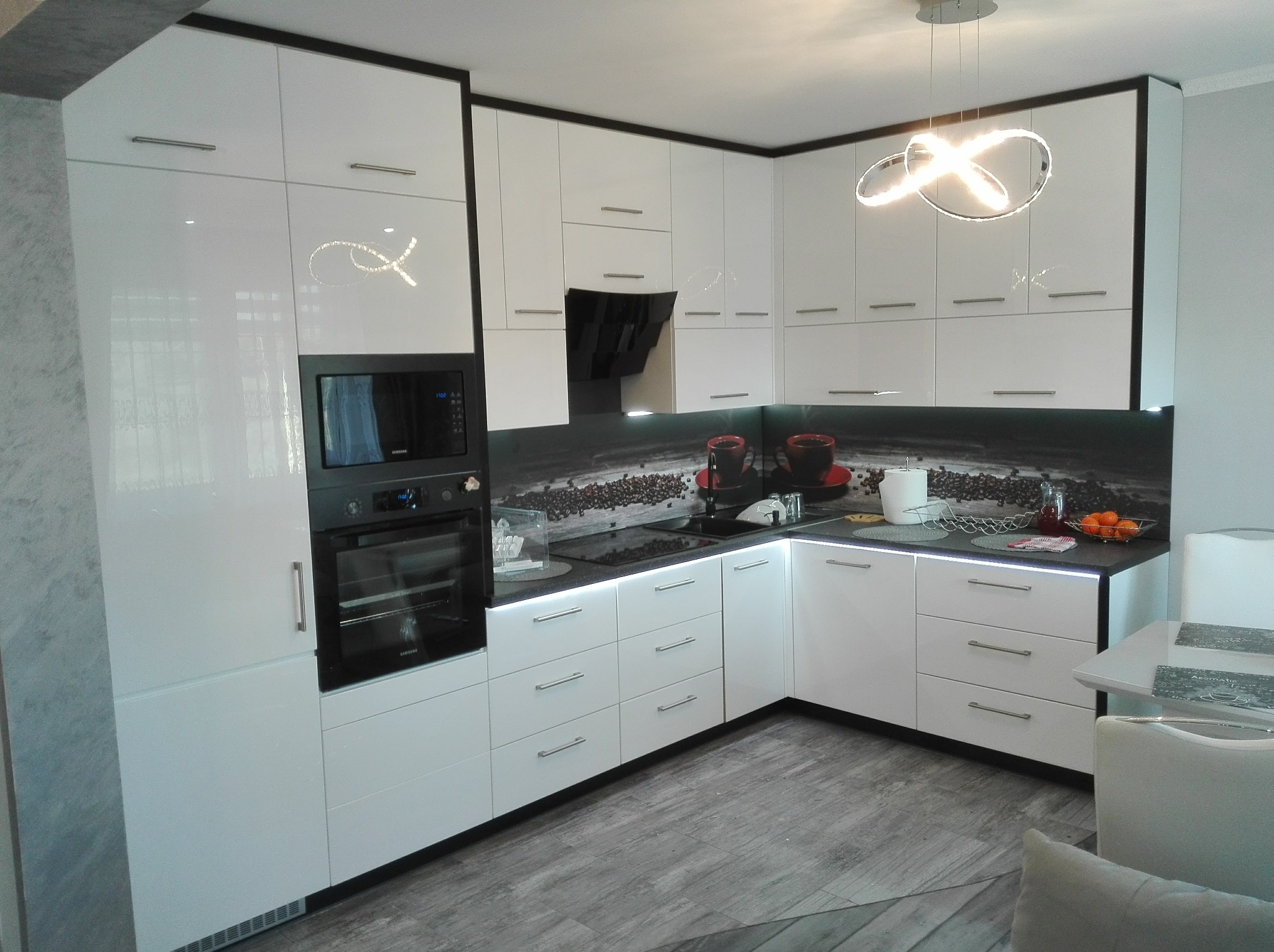 Biała, narożna kuchnia na wymiar z czarnymi akcentami i podświetleniem LED blatu. Nad blatem panel z motywem kawy. Nowoczesny design z piekarnikiem w zabudowie.