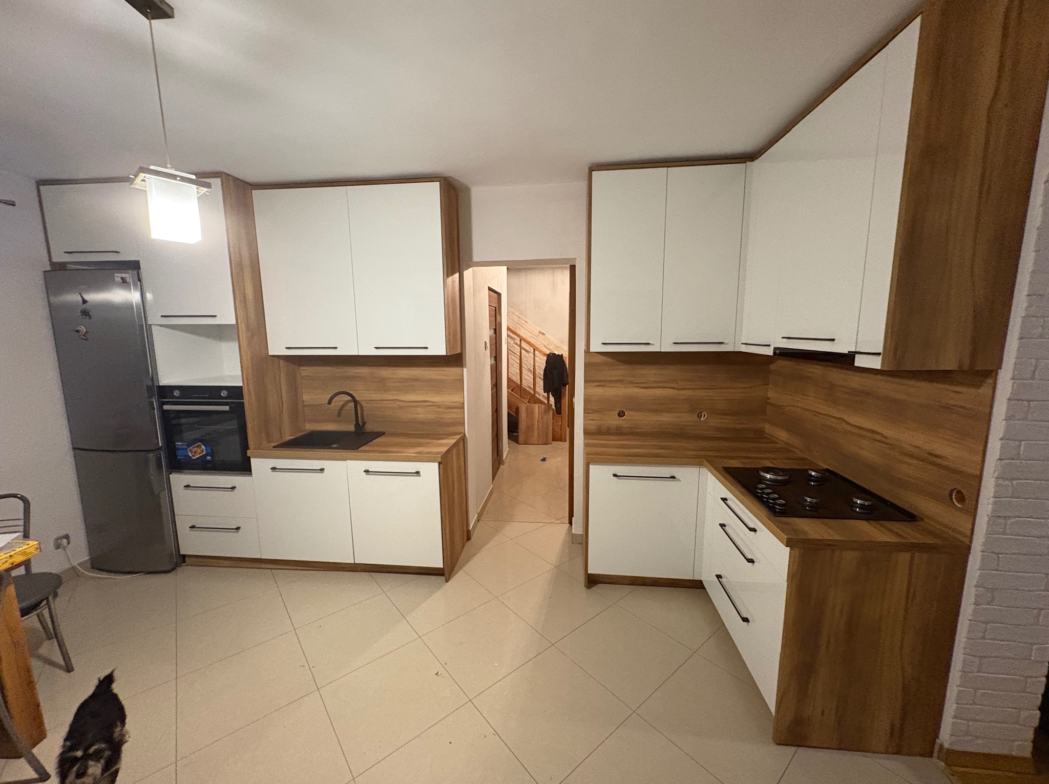 Nowoczesna kuchnia z białymi frontami i drewnianymi blatami. Widoczny zlew, płyta grzewcza i lodówka. W tle schody. Na podłodze płytki. W rogu kadru pies.
