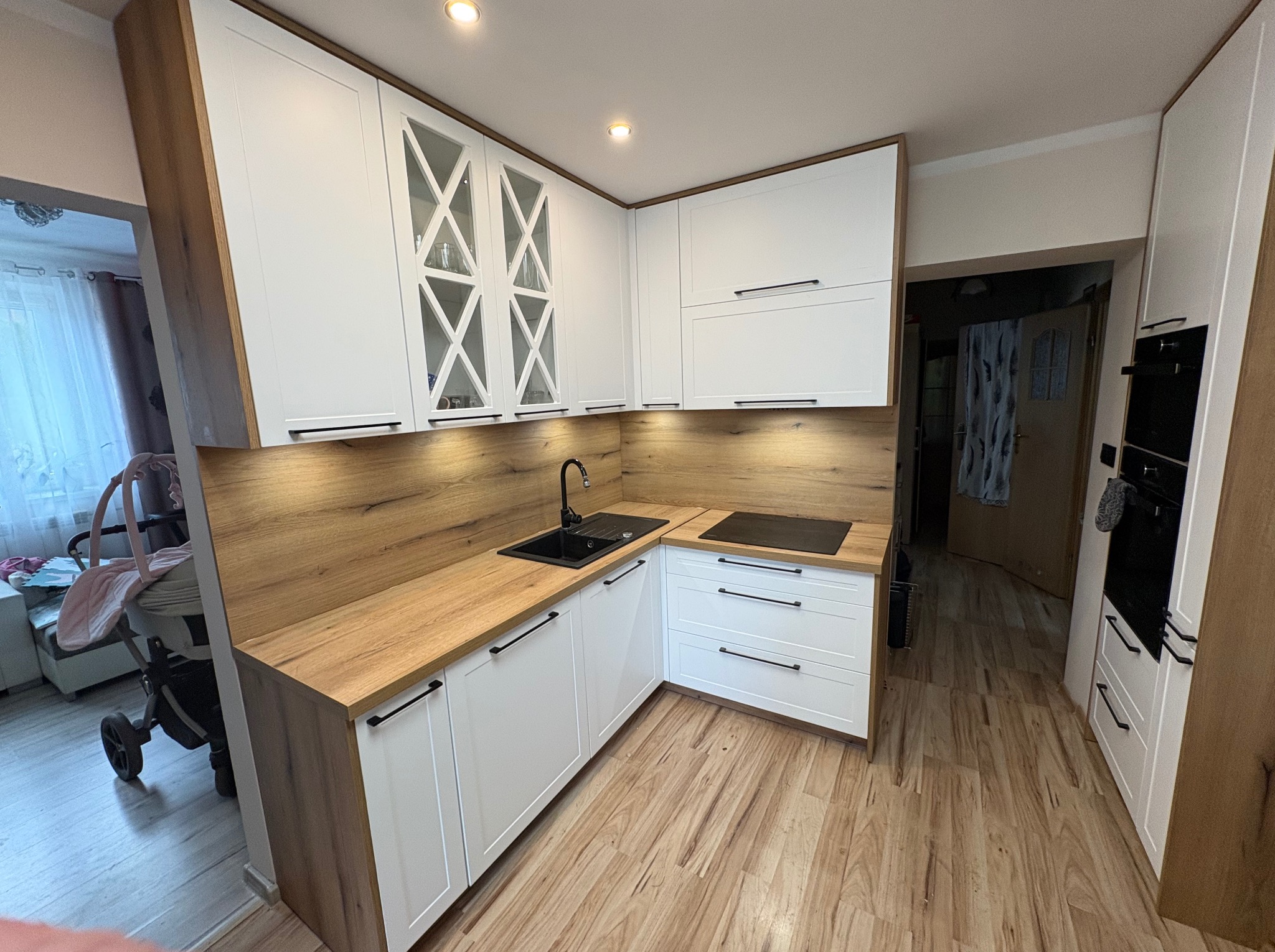 Biała kuchnia narożna z drewnianym blatem i frontami z czarnymi uchwytami. Górne szafki z przeszkleniami w kształcie krzyży. Widok na wózek dziecięcy w tle.
