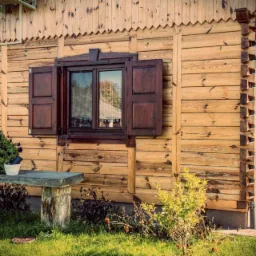 Drewniany dom z bali z otwartymi, ciemnobrązowymi okiennicami, na parapecie kwiaty w doniczce, obok ławeczka z pnia drzewa, widok na elewację i fragment werandy.