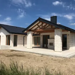 Nowoczesny dom jednorodzinny z białej cegły z zadaszonym tarasem i czarnym dachem na tle błękitnego nieba z chmurami.