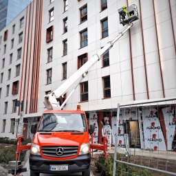 Royal Serwis Sebastian Pachocki - Czerwony podnośnik koszowy Mercedes na tle budynku z pracownikiem w koszu, wykonującym prace na wysokości. Widoczne rusztowania i elementy zabezpieczeń.