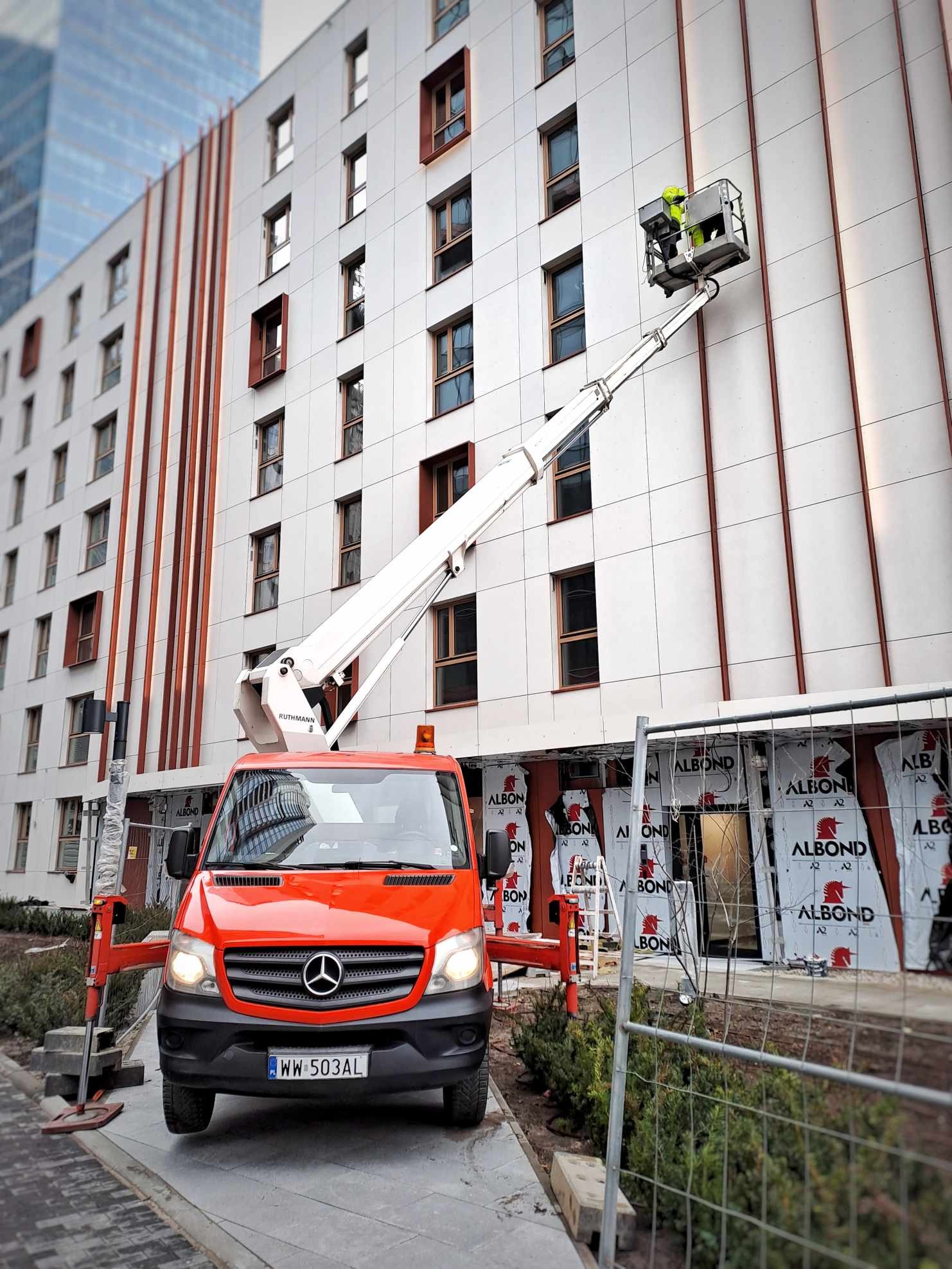 Czerwony podnośnik koszowy Mercedes na tle budynku z pracownikiem w koszu, wykonującym prace na wysokości. Widoczne rusztowania i elementy zabezpieczeń.