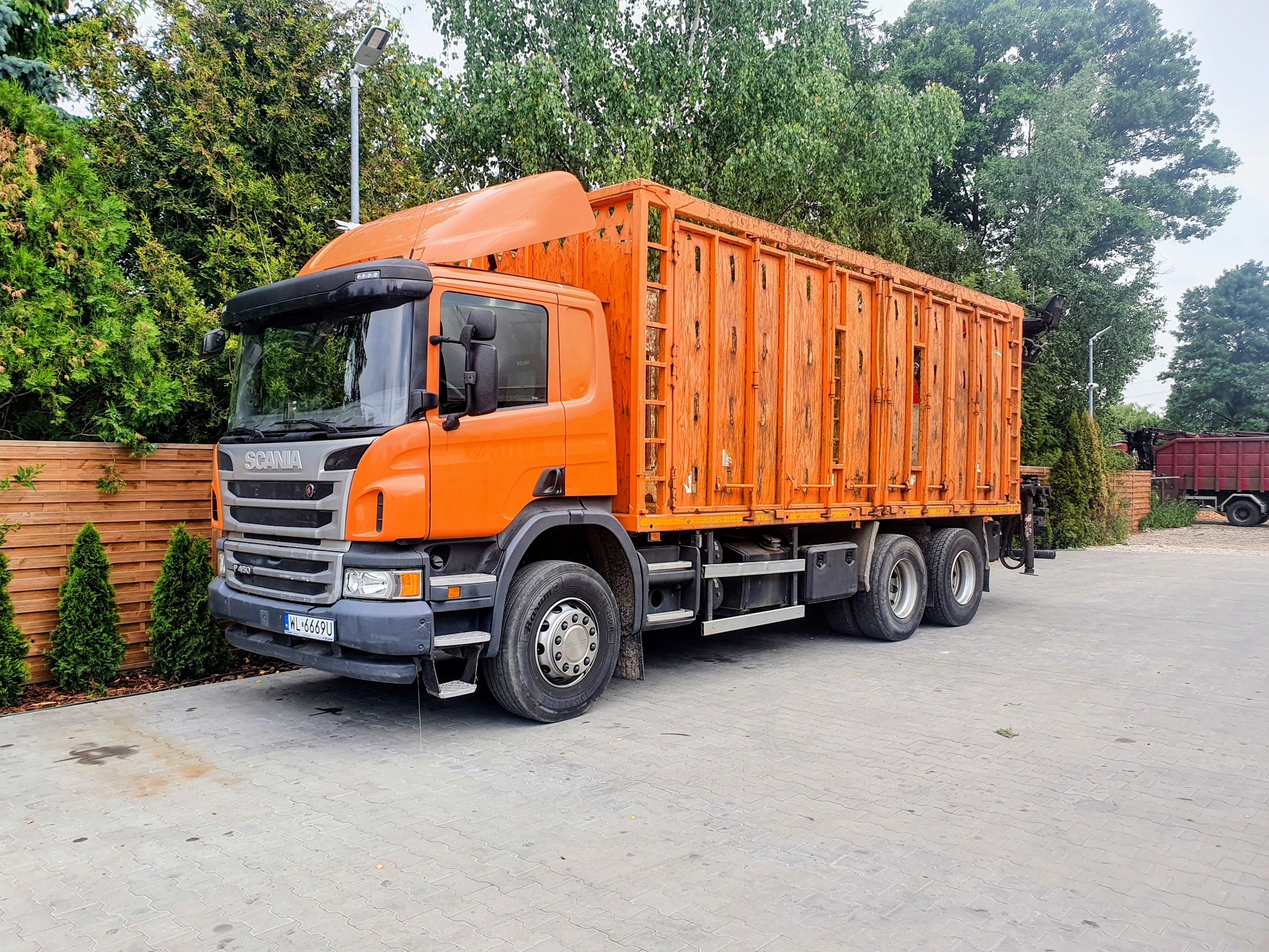 Pomarańczowa ciężarówka Scania z ażurową zabudową skrzyniową na tle zieleni. Widoczny fragment ogrodzenia i oświetlenie uliczne. Tablice rejestracyjne z przodu pojazdu.