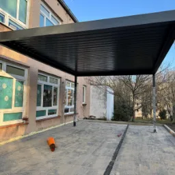 Ciemna, nowoczesna pergola z metalu przylega do budynku. Słupy częściowo owinięte folią zabezpieczającą, pomarańczowy pachołek leży na szarym bruku.
