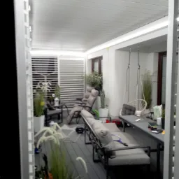 Nowoczesna, zadaszona przestrzeń tarasowa z szarym tarasem kompozytowym, białymi lamelami i meblami wypoczynkowymi. Widoczne rośliny w donicach i wiszące huśtawki.