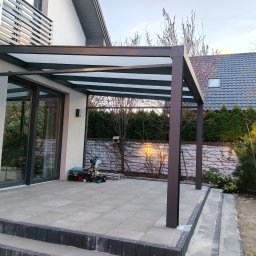 PW ARTINOX - Nowoczesna pergola metalowa z zadaszeniem z poliwęglanu, przylegająca do domu z klimatyzacją. Widoczne narzędzia na tarasie z szarej kostki.