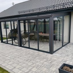 PW ARTINOX - Szklana zabudowa tarasu z czarnymi ramami w Nidzicy. Widoczny stół i krzesła wewnątrz. Eleganckie wykończenie, szara kostka brukowa i trawnik.