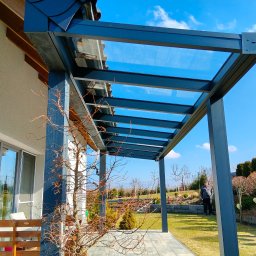 PW ARTINOX - Nowoczesna pergola metalowa z przeszklonym dachem, wsparta na słupach, przylegająca do budynku. Widoczny zadbany ogród i osoba w tle. Słoneczny dzień.