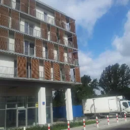 Nowoczesny budynek z balkonami i drewnianymi żaluzjami fasadowymi. Metalowe balustrady balkonowe. W tle błękitne niebo i zieleń drzew.