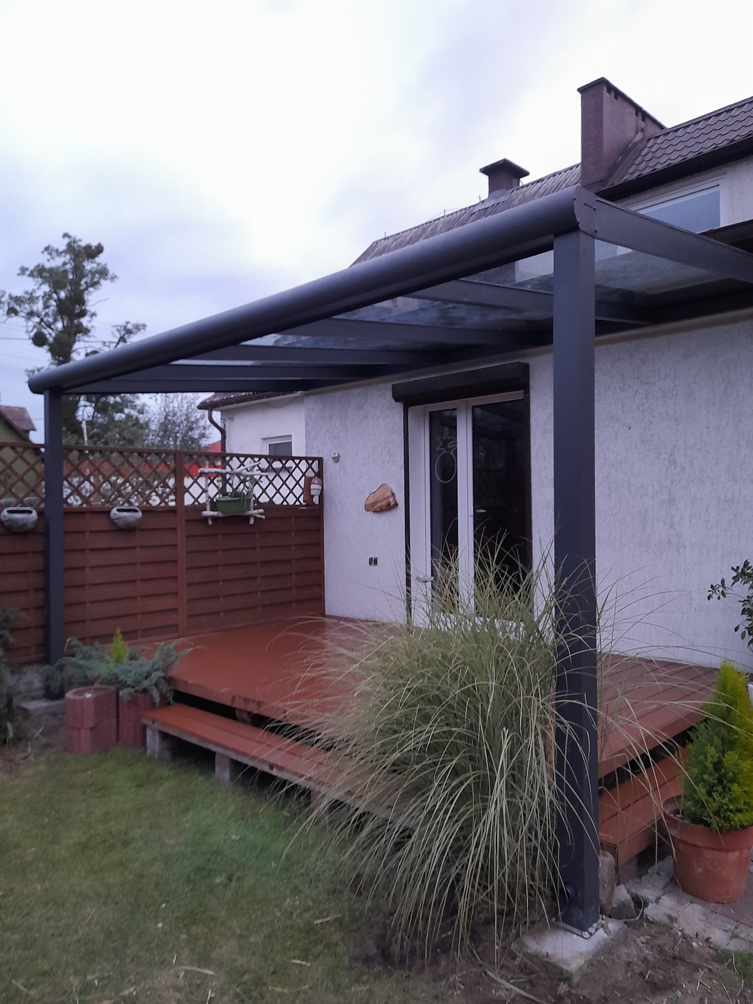 Nowoczesna pergola z metalową konstrukcją i przeszklonym dachem, osłaniająca drewniany taras z niskimi schodkami i zielenią wokół.