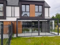 Nowoczesna, przeszklona zabudowa tarasu z ciemną ramą, przylegająca do jasnego domu z drewnianymi akcentami. Widoczny fragment metalowego ogrodzenia i trawnik.