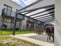 Nowoczesna pergola z szarymi słupami i białym zadaszeniem na tarasie z jasnych płyt. W tle budynek mieszkalny i zielony trawnik.