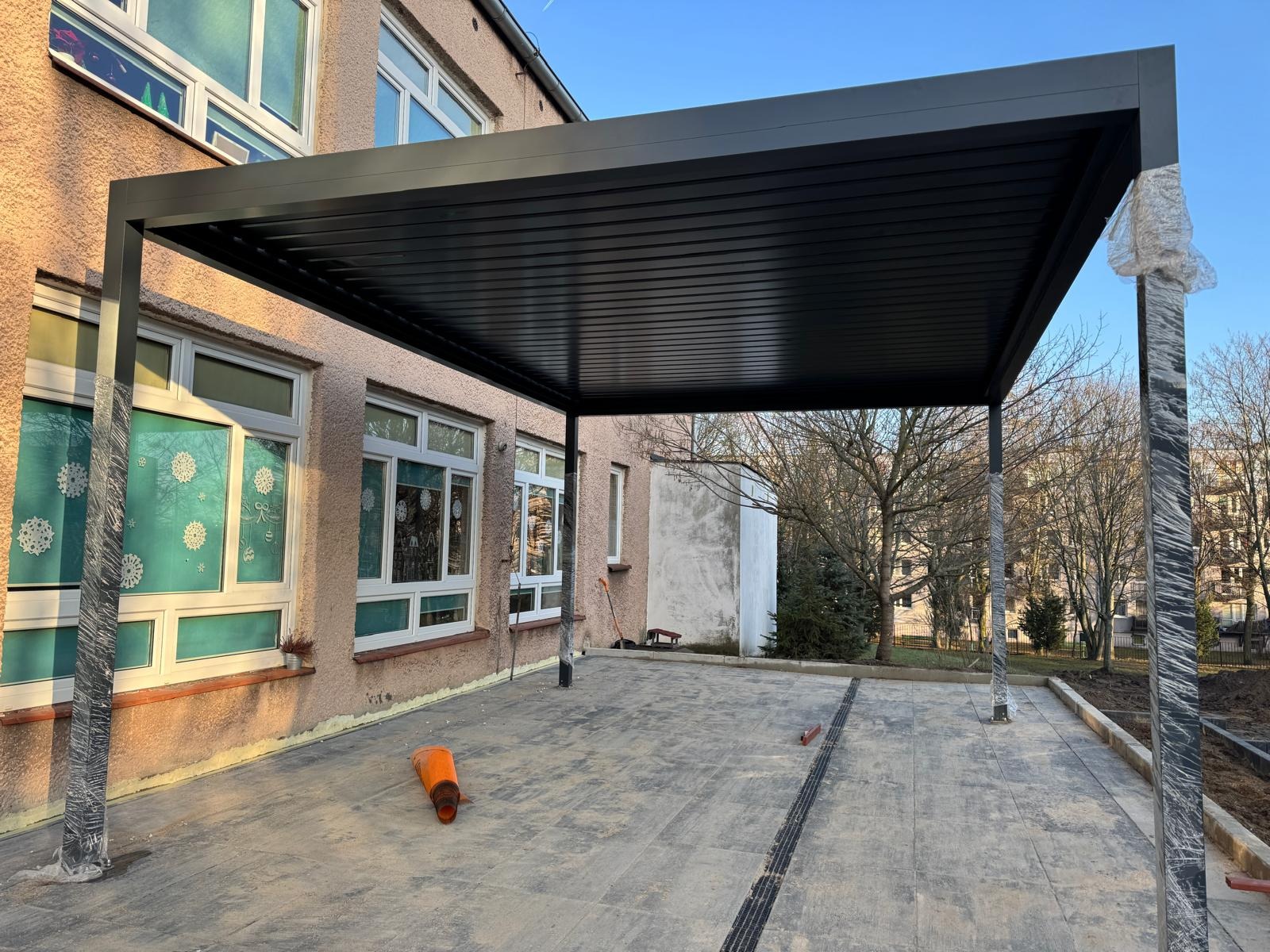 Ciemna, nowoczesna pergola z metalu przylega do budynku. Słupy częściowo owinięte folią zabezpieczającą, pomarańczowy pachołek leży na szarym bruku.