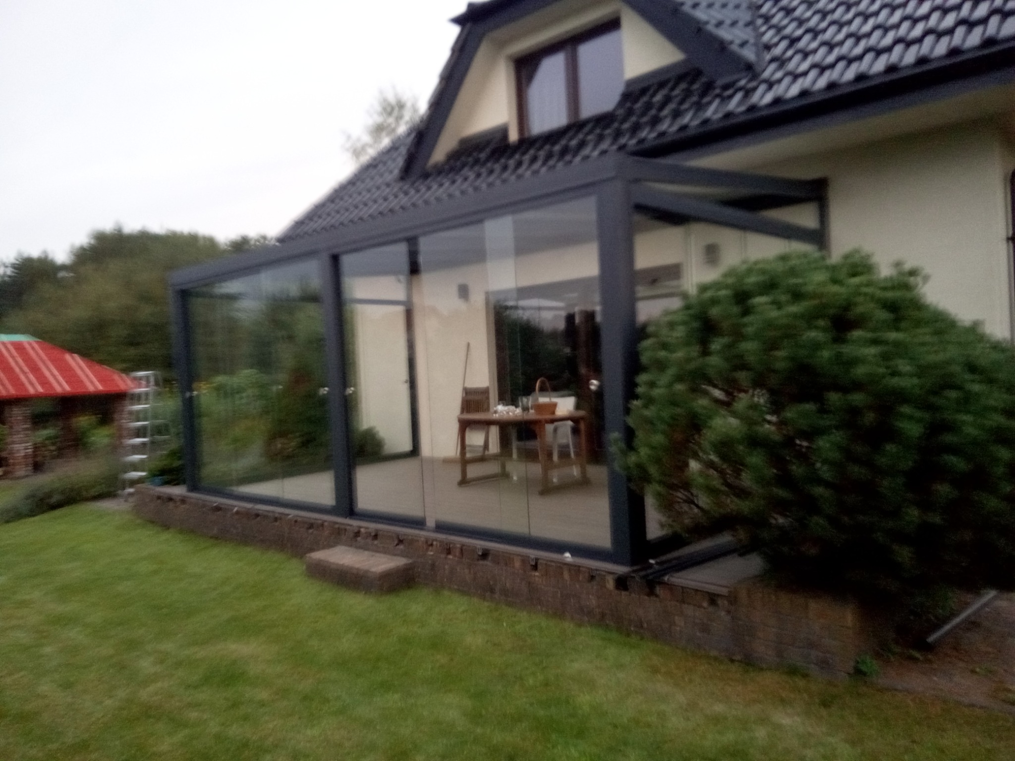 Szklana zabudowa tarasu z ciemną ramą, widok z zewnątrz na trawnik i fragment domu. Wewnątrz widoczny stół i krzesła. Na zewnątrz rośnie duży krzew.