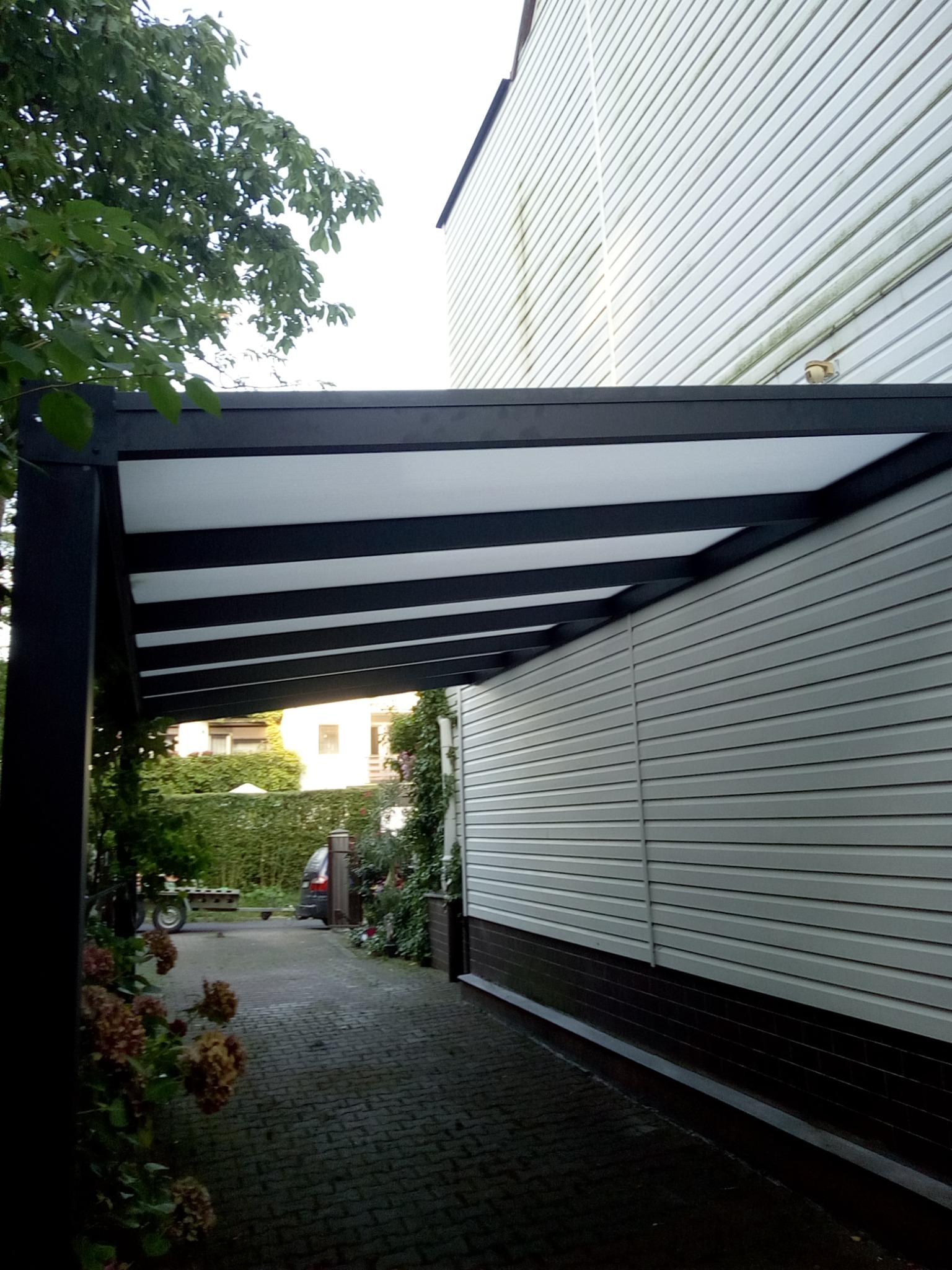 Nowoczesna pergola metalowa z białym zadaszeniem, przylegająca do budynku z sidingiem. Widok na brukowaną ścieżkę i zieleń. Minimalistyczny design, czarne elementy konstrukcyjne.