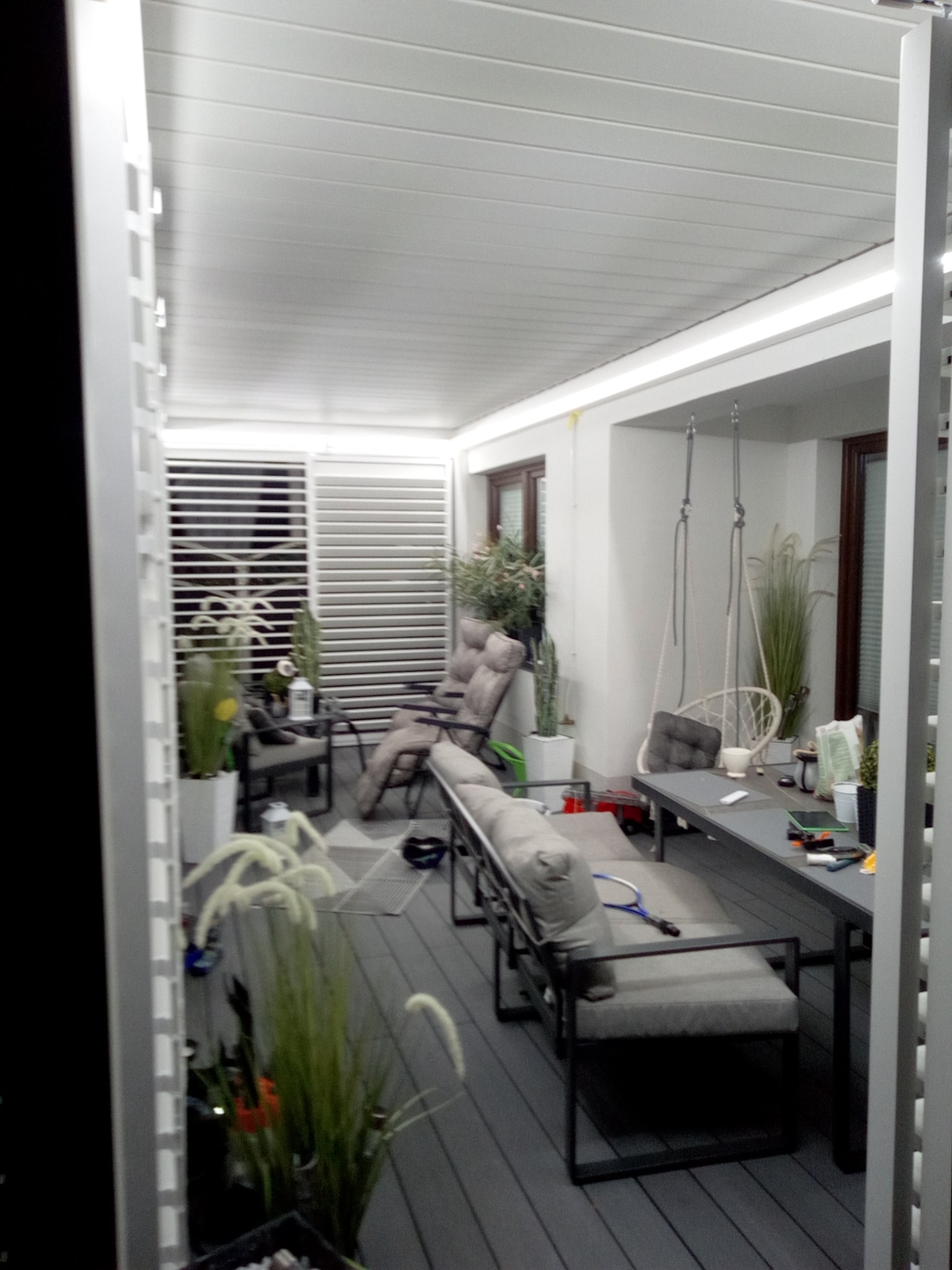 Nowoczesna, zadaszona przestrzeń tarasowa z szarym tarasem kompozytowym, białymi lamelami i meblami wypoczynkowymi. Widoczne rośliny w donicach i wiszące huśtawki.