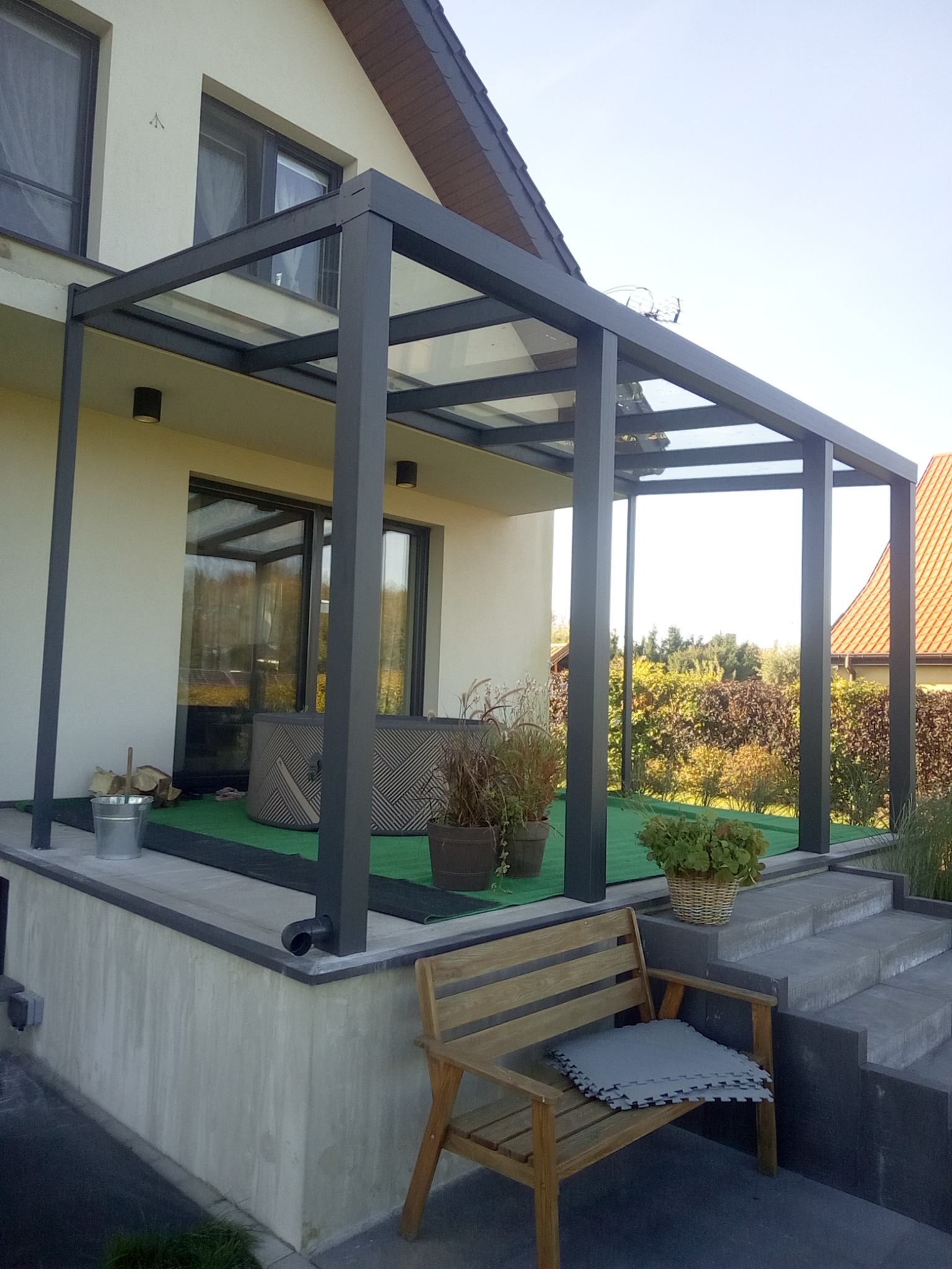 Nowoczesna pergola metalowa z przezroczystym zadaszeniem, przylegająca do jasnego budynku. Widoczne schody, rośliny w donicach i drewniana ławka.