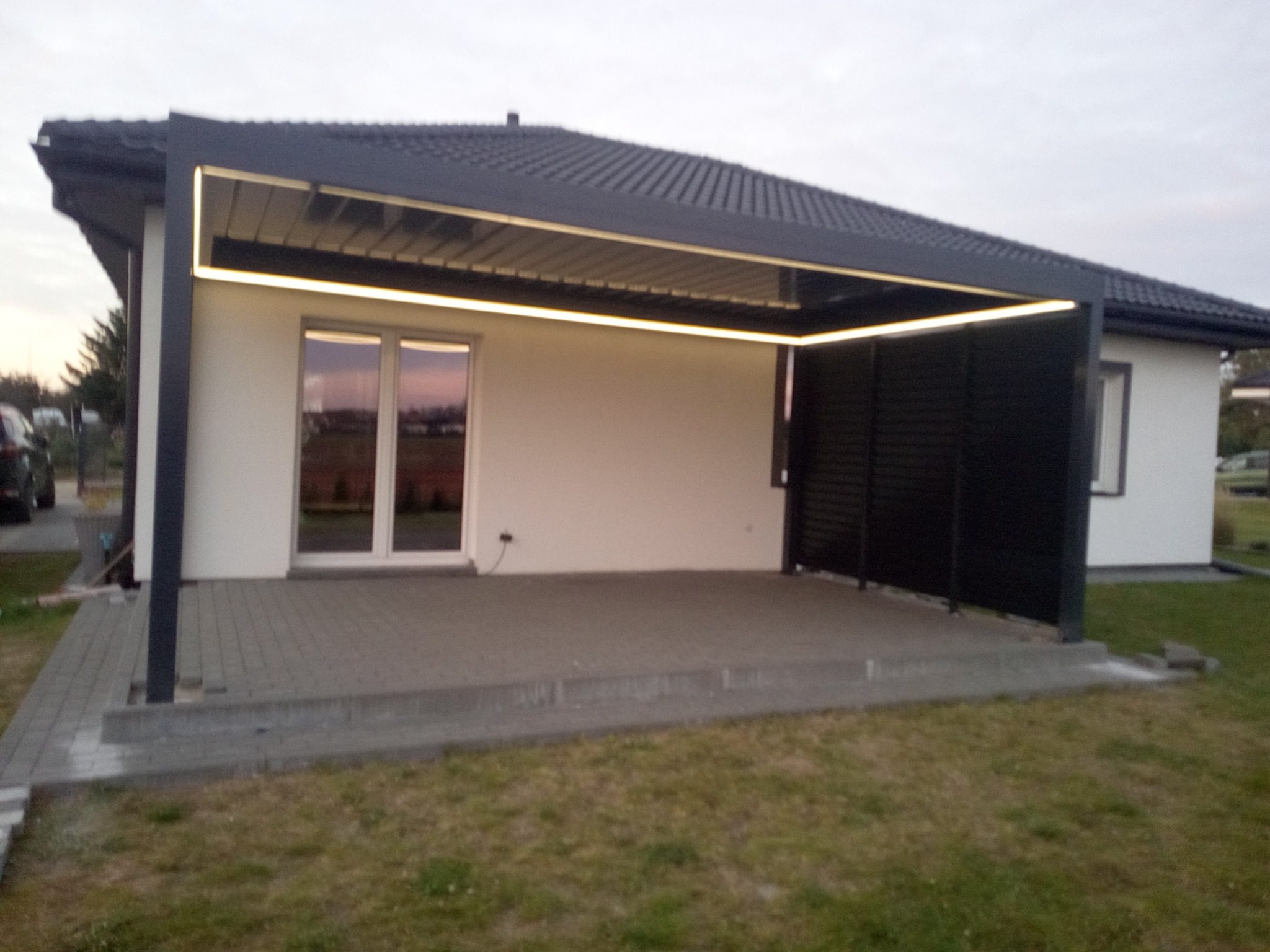 Nowoczesna, czarna pergola metalowa z oświetleniem LED, przylegająca do białego budynku. Widoczny fragment tarasu z kostki brukowej i trawnik.