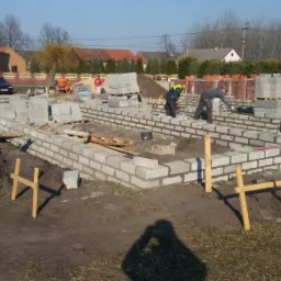 Budowa fundamentów z bloczków betonowych w Stopnicy, z widocznymi pracownikami murującymi ściany i stertami materiałów budowlanych w tle.