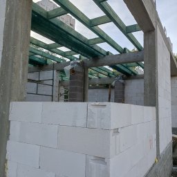 Zakład budowlany - Budowa domu w stanie surowym otwartym: białe bloczki, betonowe słupy i zielona więźba dachowa. Widoczne kominy i rusztowania wewnątrz budynku.