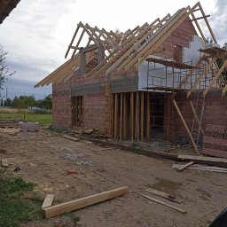 Alekbud - Budowa domu z czerwonej cegły w trakcie wznoszenia więźby dachowej. Widoczne rusztowania i materiały budowlane. Ujęcie z poziomu gruntu, pochmurne niebo w tle.