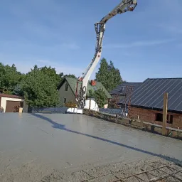 Wylewanie betonu na strop zbrojony, widoczna pompa do betonu. W tle domy jednorodzinne i błękitne niebo. Prace budowlane w toku, etap zalewania stropu.