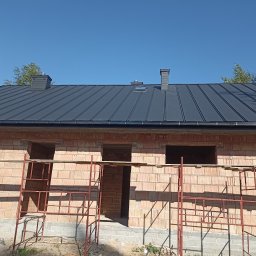 Alekbud - Budowa domu z czerwonej cegły z zamontowanym, ciemnym dachem z blachy trapezowej. Widoczne rusztowania i okna bez szyb. Błękitne niebo w tle.