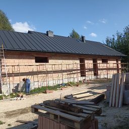 Alekbud - Budowa domu z czerwonej cegły, czarny dach, rusztowania, pracownik w niebieskim kombinezonie przy fundamentach. Widok na plac budowy w słoneczny dzień.