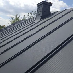 Szary dach z blachy trapezowej z kominem na tle błękitnego nieba z chmurami. Widok z dołu, ukazujący strukturę blachy i detale komina.