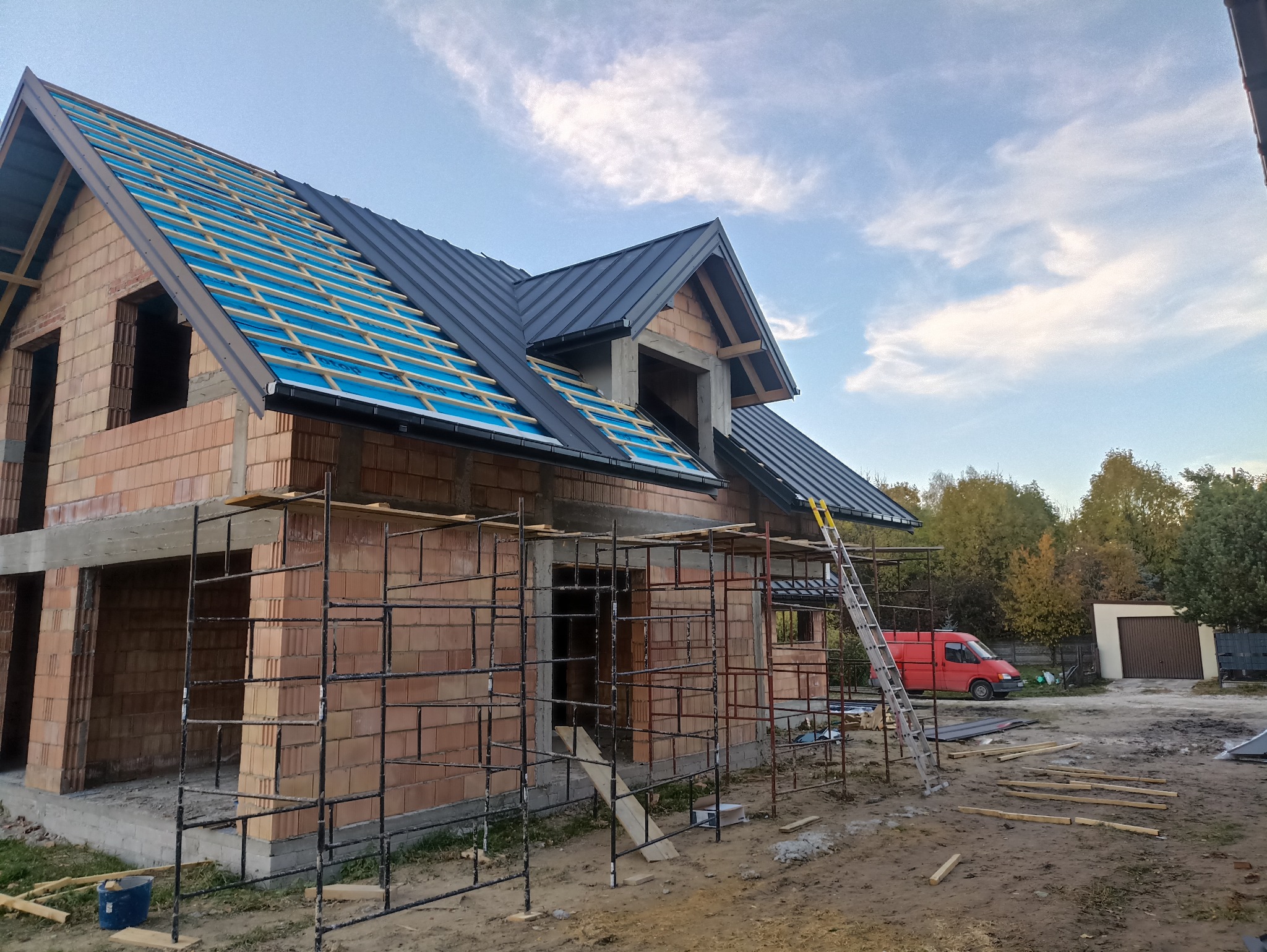 Budowa domu z czerwonej cegły. Widoczna więźba dachowa z czarnym pokryciem i niebieską membraną. Na pierwszym planie rusztowania i drabina. W tle czerwony samochód dostawczy.