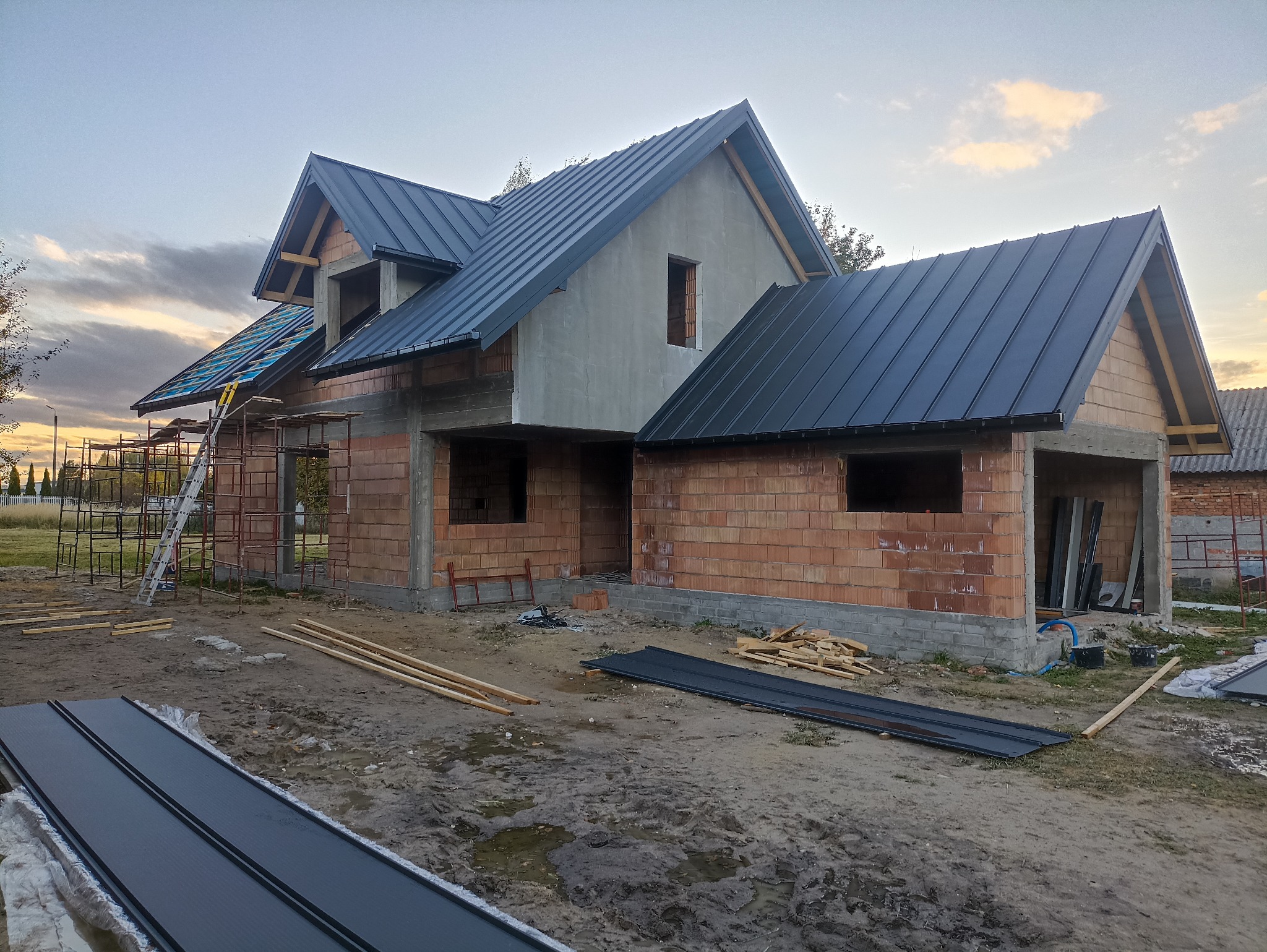 Budowa domu jednorodzinnego w trakcie realizacji: ściany z czerwonej cegły, częściowo pokryty ciemną blachą dach, rusztowania, elementy więźby dachowej. Widok z zewnątrz.