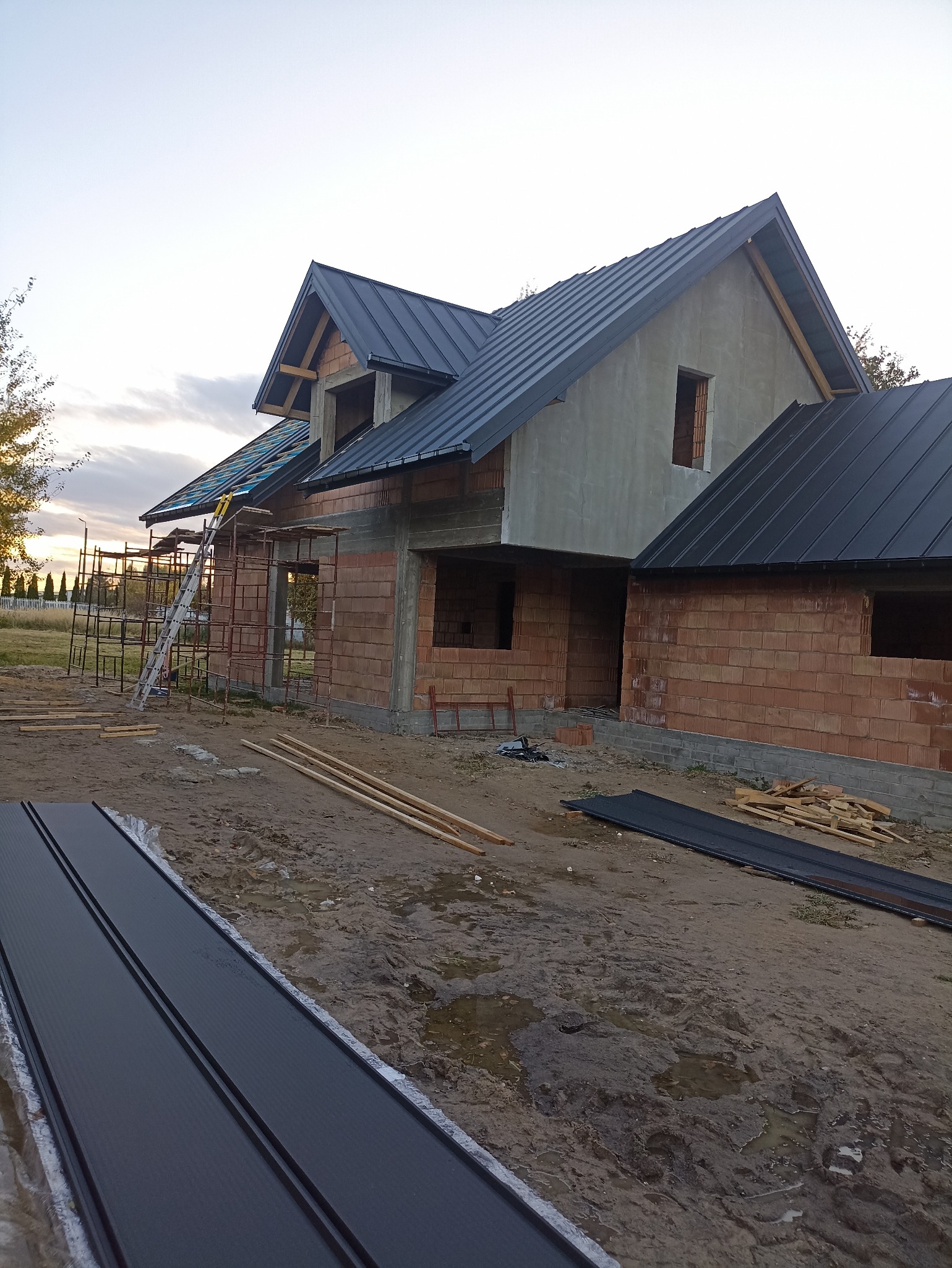 Budowa domu w trakcie realizacji: mury z czerwonej cegły, częściowo pokryty ciemną blachą dach, rusztowania i materiały budowlane na placu. Widok z dołu.