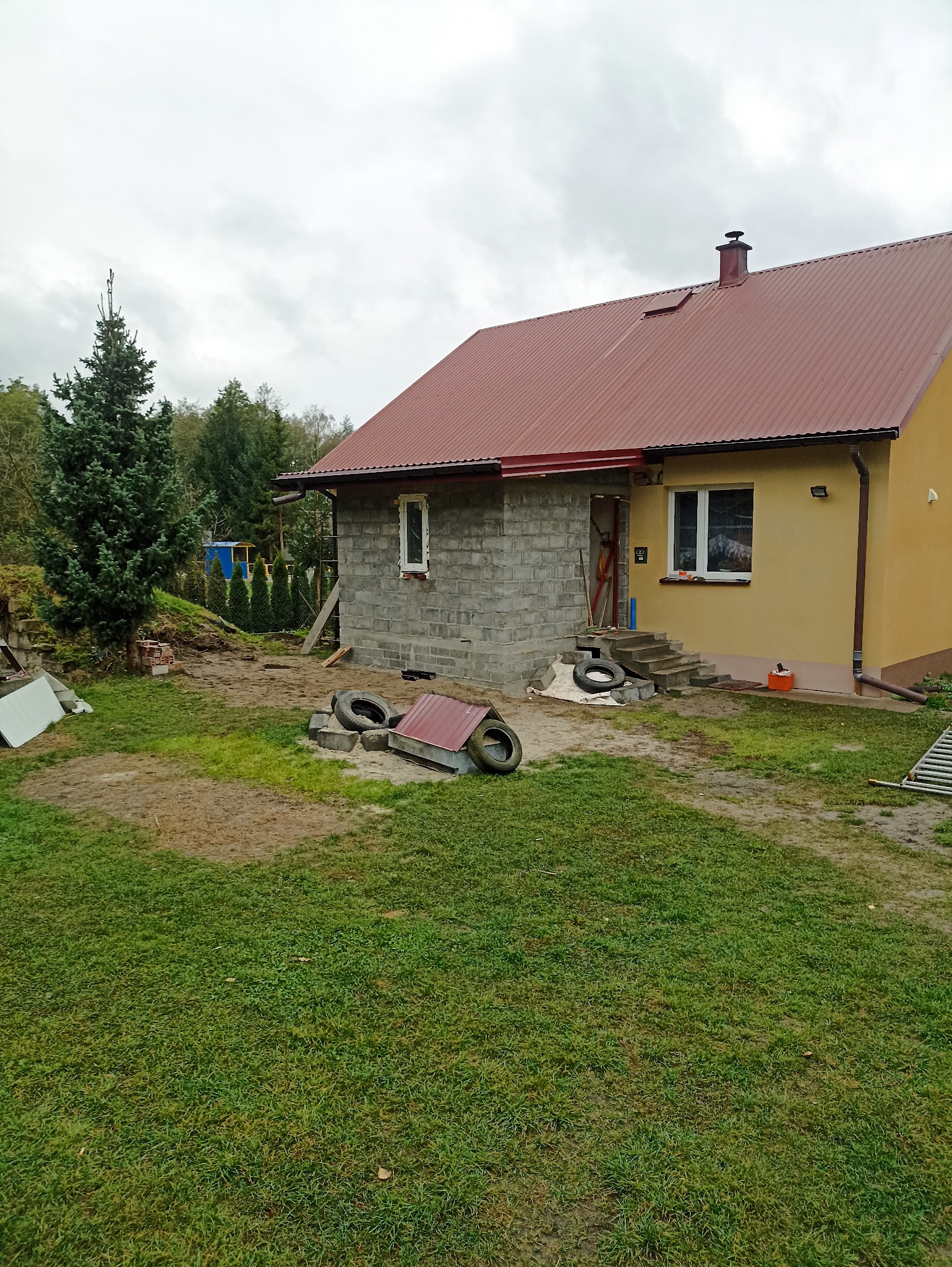 Rozbudowa domu z czerwonym dachem: dobudowana część z szarego bloczka, widoczne opony i gruz na trawniku. Część domu pomalowana na żółto.