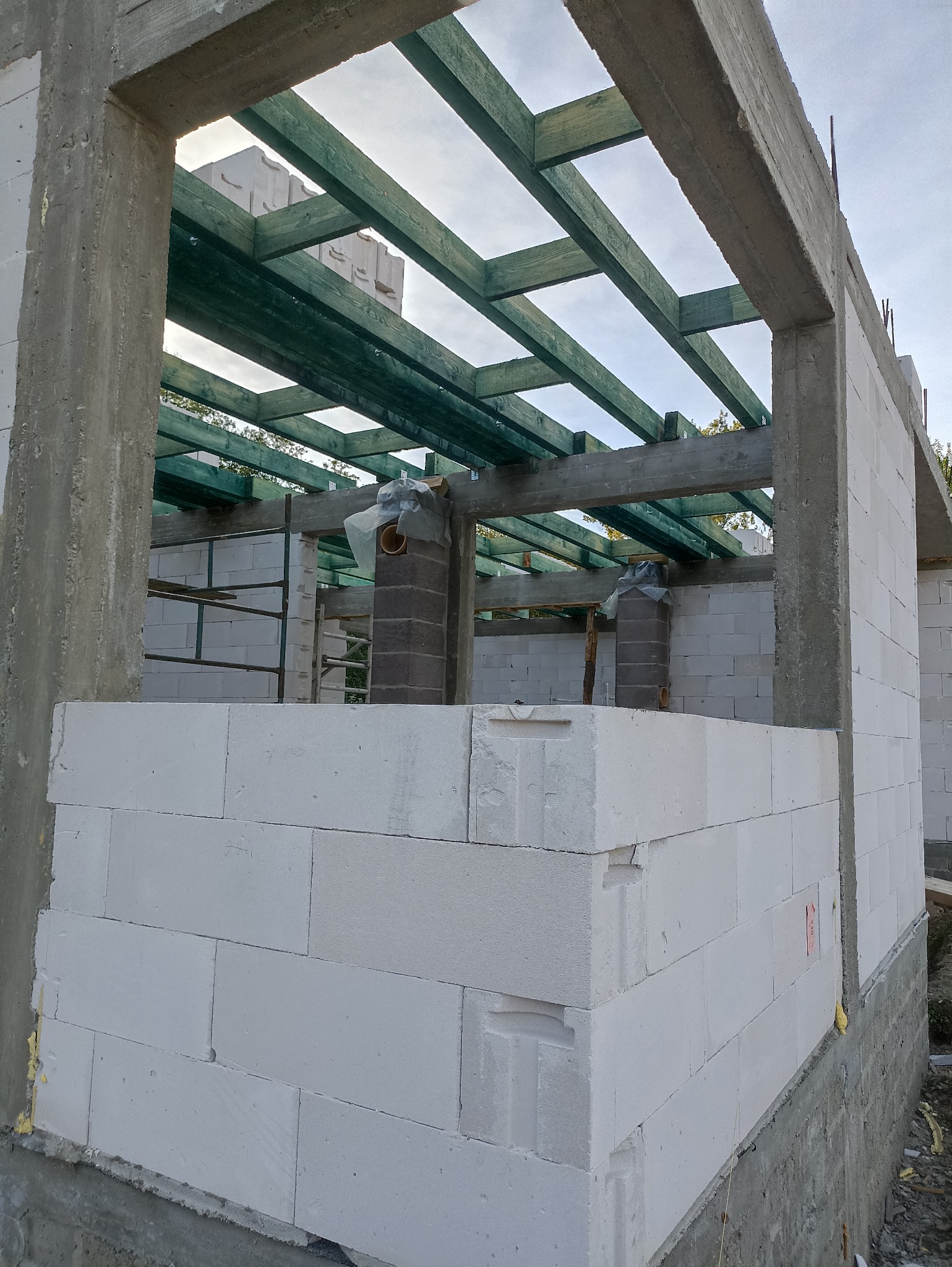 Budowa domu w stanie surowym otwartym: białe bloczki, betonowe słupy i zielona więźba dachowa. Widoczne kominy i rusztowania wewnątrz budynku.