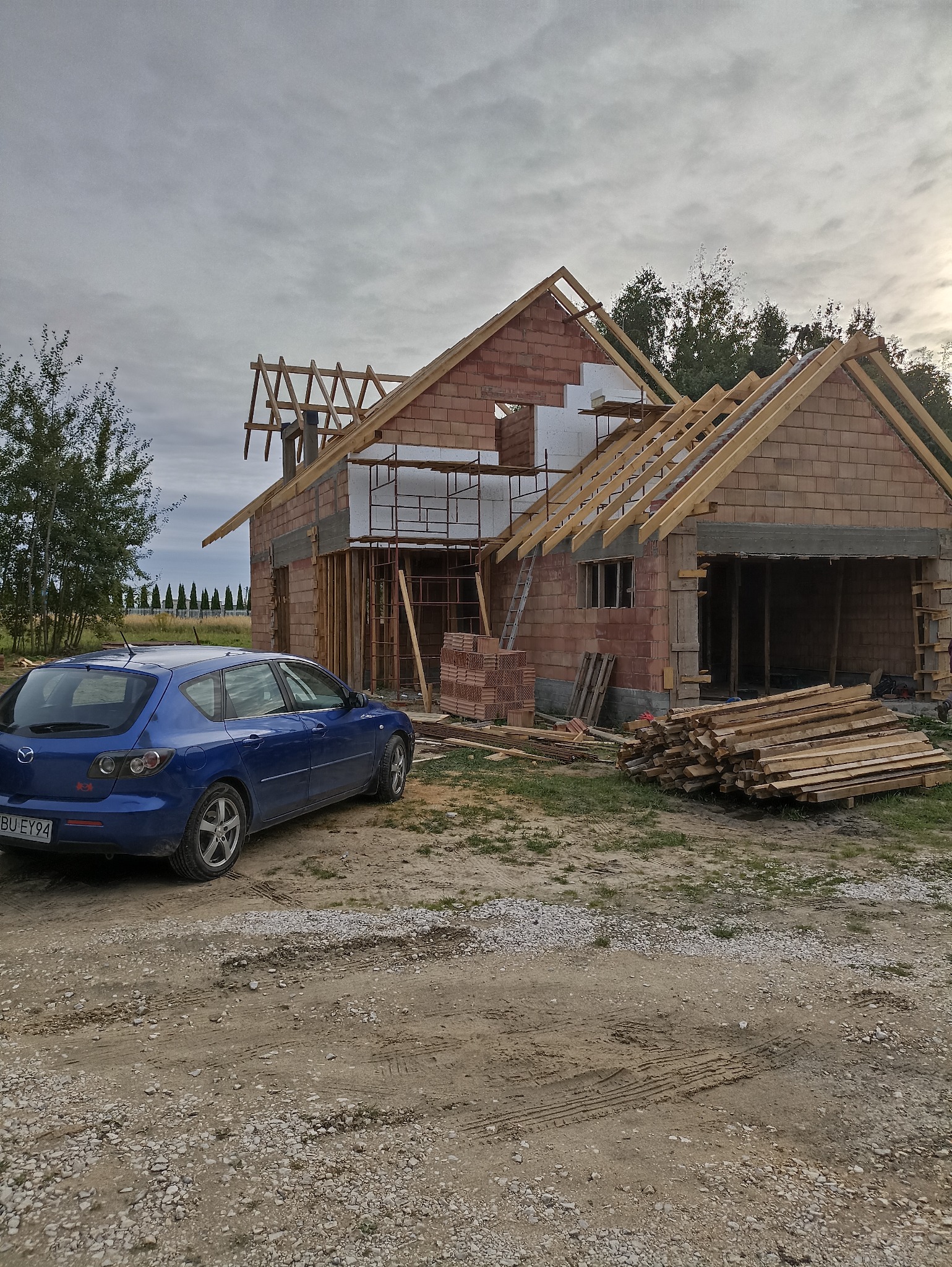 Surowy stan domu z czerwonej cegły w trakcie budowy, z widoczną więźbą dachową i częściowo ocieplonymi ścianami. Na pierwszym planie zaparkowany niebieski samochód.
