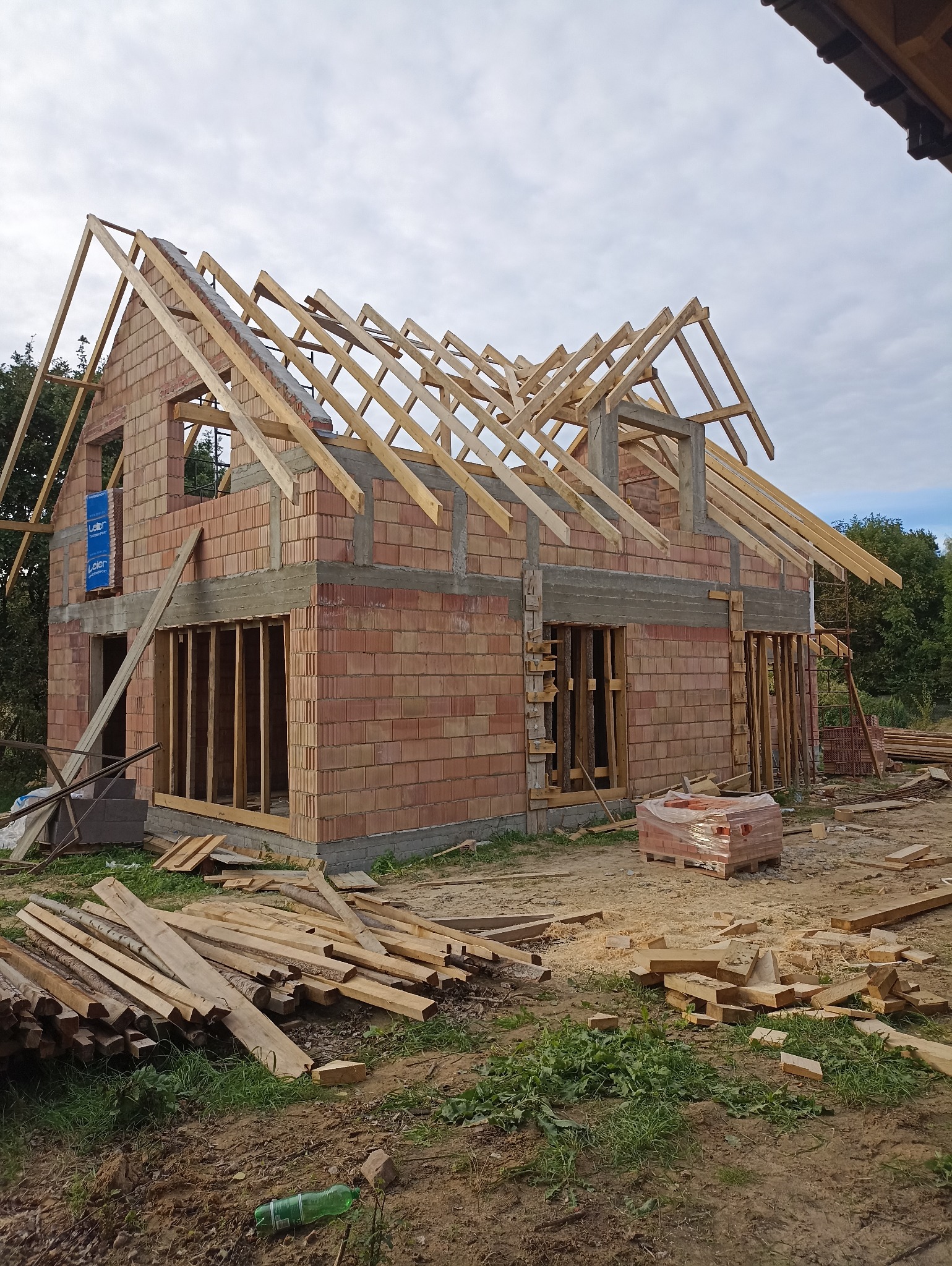 Surowy stan domu z czerwonej cegły z drewnianą konstrukcją dachu w trakcie budowy. Widoczne materiały budowlane i otoczenie placu budowy.