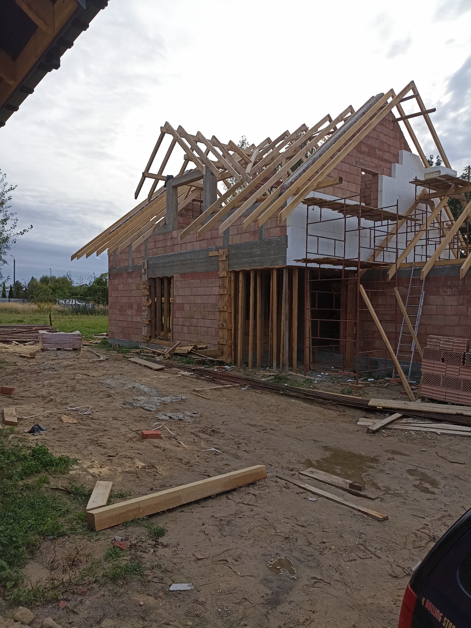 Budowa domu z czerwonej cegły w trakcie wznoszenia więźby dachowej. Widoczne rusztowania i materiały budowlane. Ujęcie z poziomu gruntu, pochmurne niebo w tle.